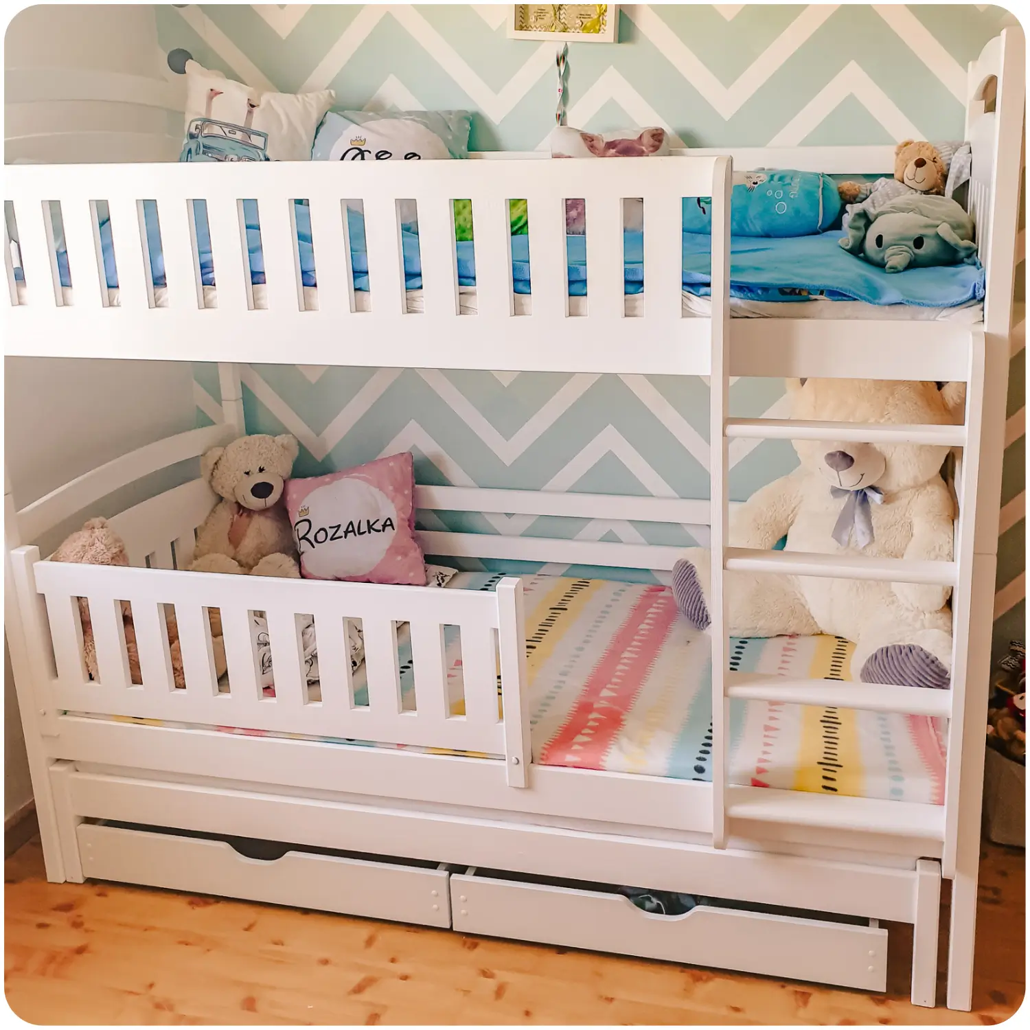 White children's bunk bed BLANKA for three people, with a pull-out bottom bed. It has high protective railings on both levels, a vertical ladder, and two pull-out drawers. Placed against a wall with turquoise and white zigzag wallpaper.