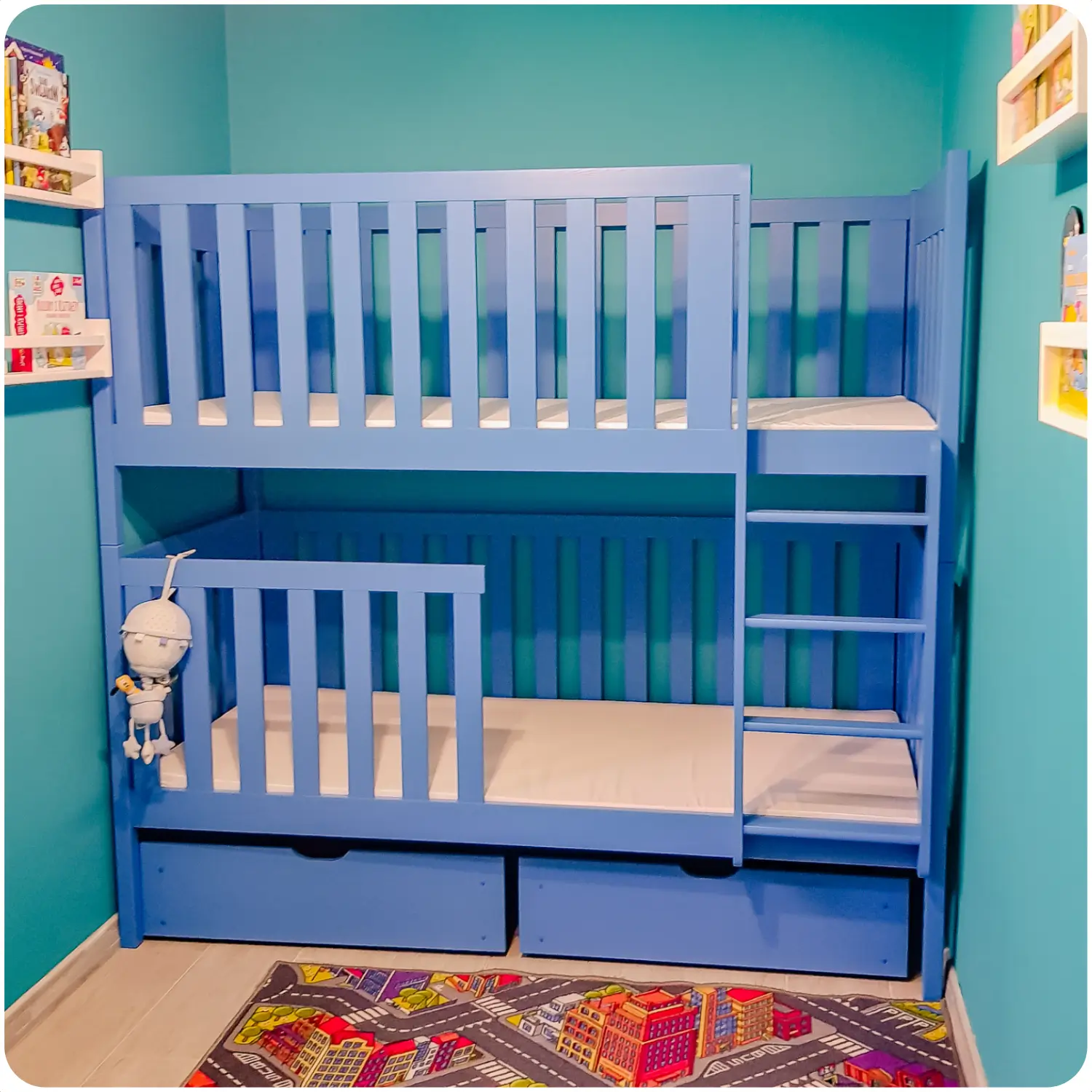 Navy blue bunk bed for children with pull-out drawers. Equipped with high railings and a vertical ladder. The room has intensely blue walls, a light floor, and a carpet with a colorful street pattern. Shelves with books and toys hang on the walls.