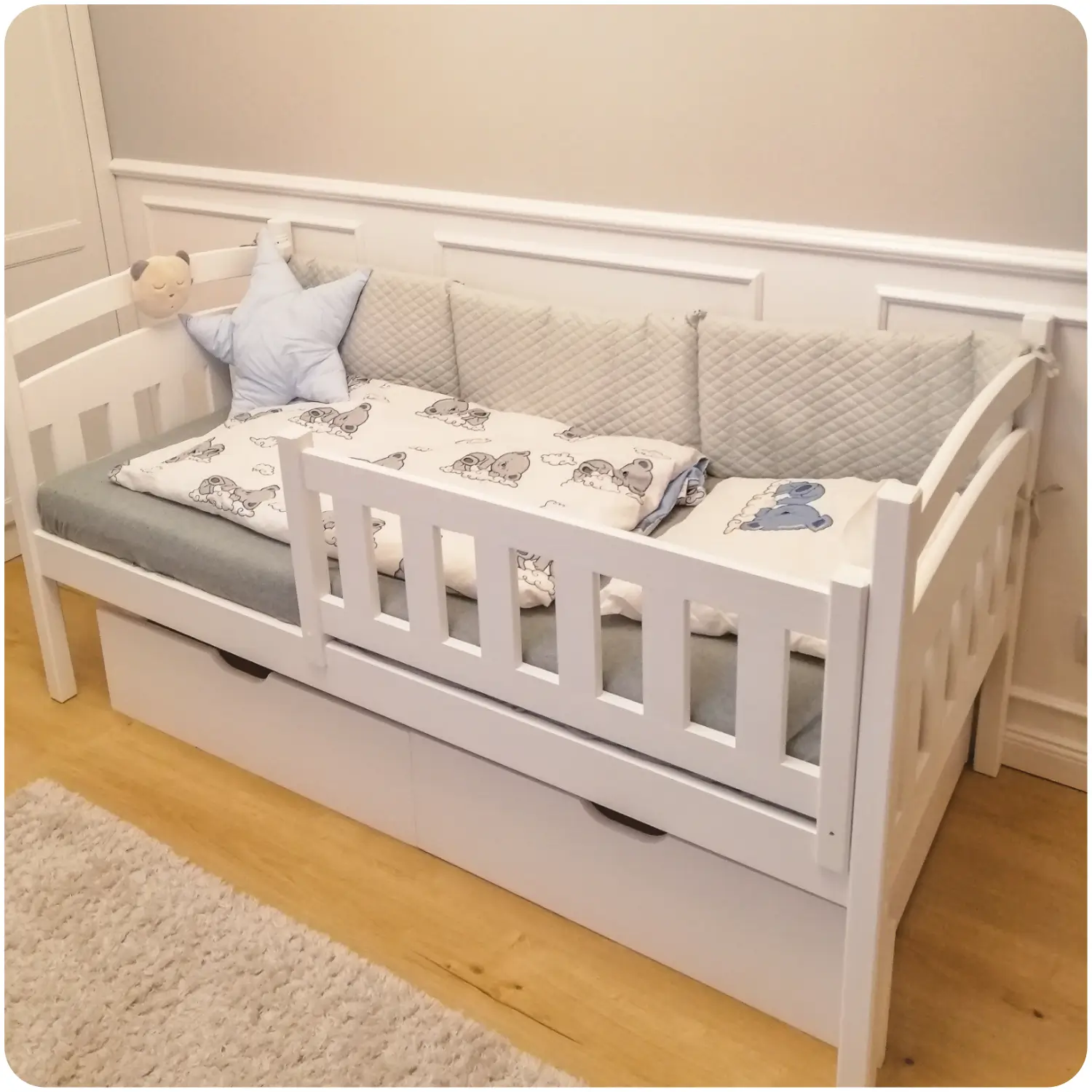 White single children's bed with rounded headboards and a removable protective railing. Equipped with two drawers for bedding or toys. Decorated in a subdued beige color scheme with grey bumpers and white bedding with bears.